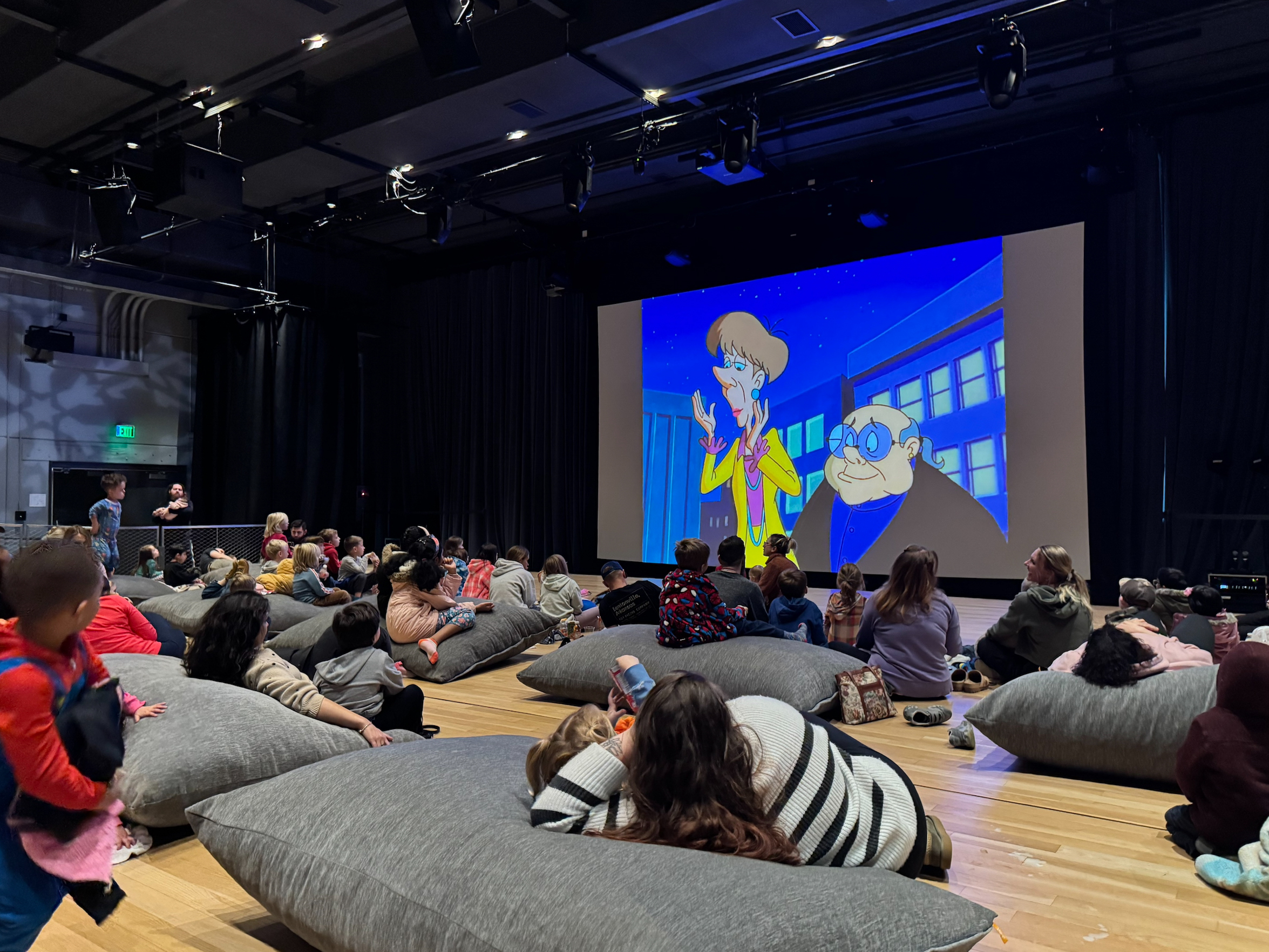 Children and adults seated on gray cushions watching a cartoon on a large screen in a dark room.