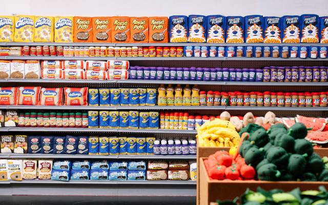 Supermarket aisle with shelves of packaged foods and a box of plush fruits and vegetables.