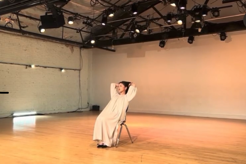 Person in a light-colored dress sitting on a chair in a large, well-lit room with wooden floor.