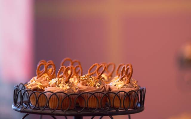 Cupcakes with light brown frosting, gold foil, and mini pretzels on a black metal stand.