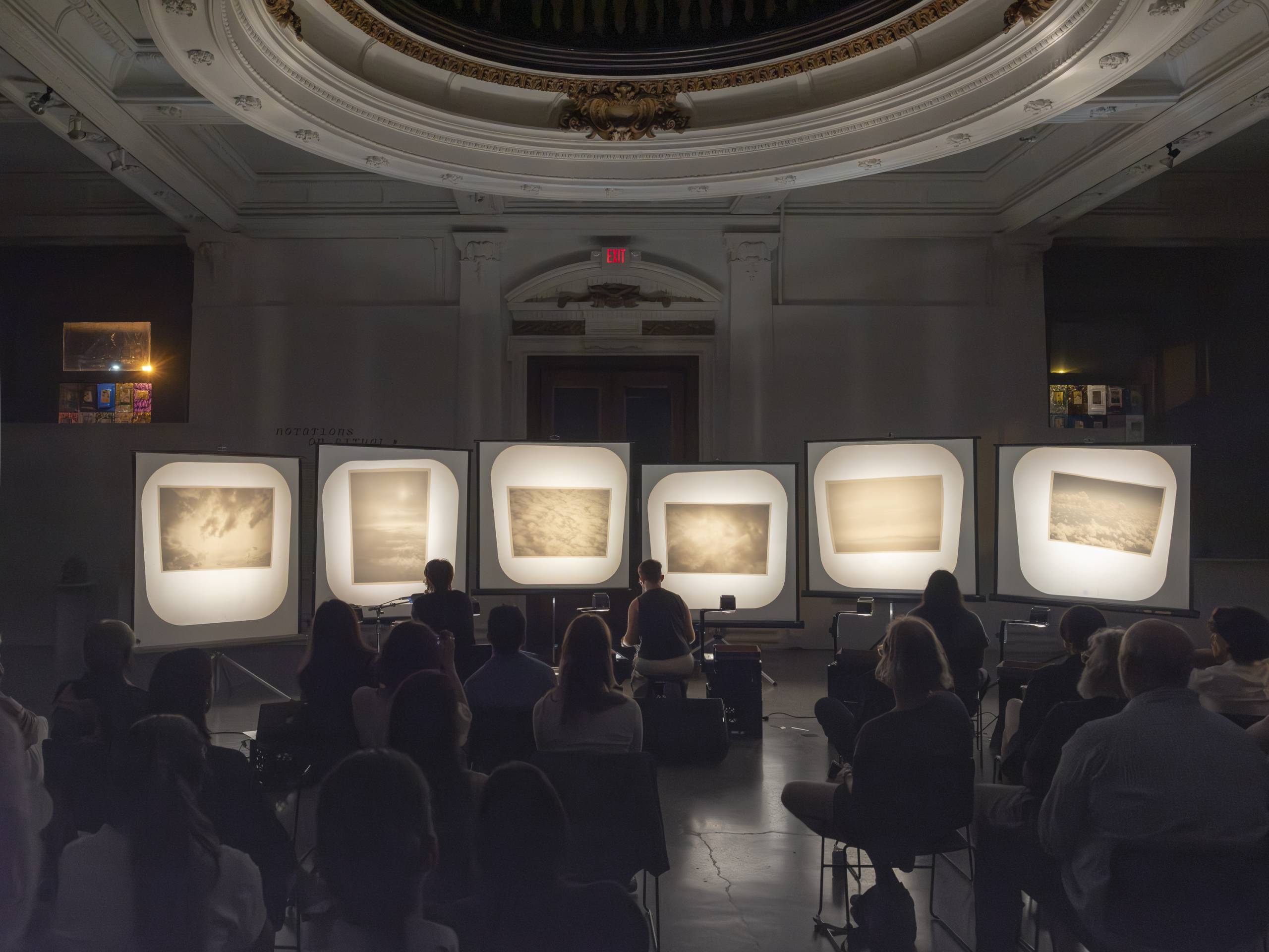 Audience seated in a dim room watching five illuminated screens showing cloud images in a decorative hall.