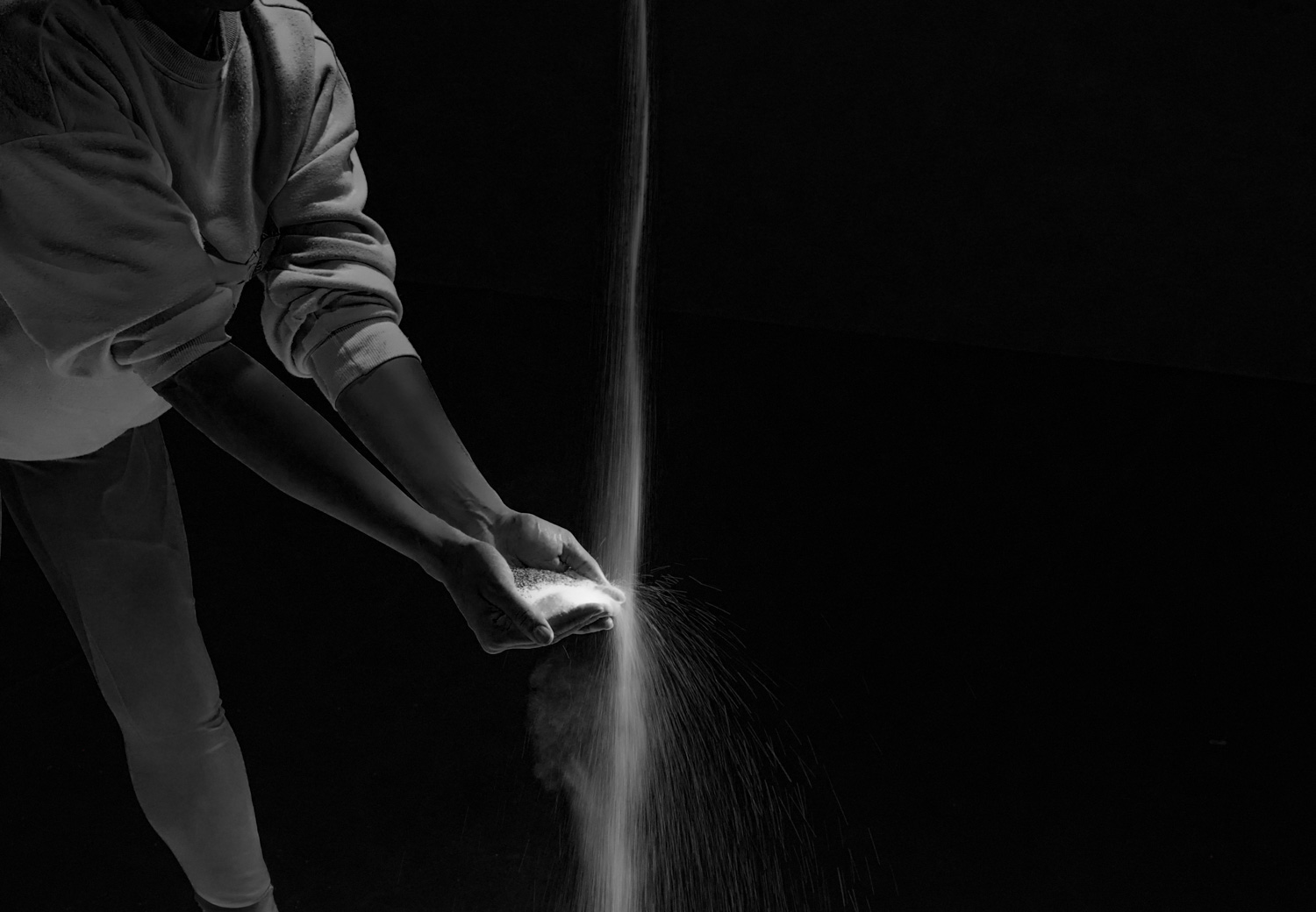 Person in long-sleeve shirt pouring a fine powder or sand from cupped hands over a dark background