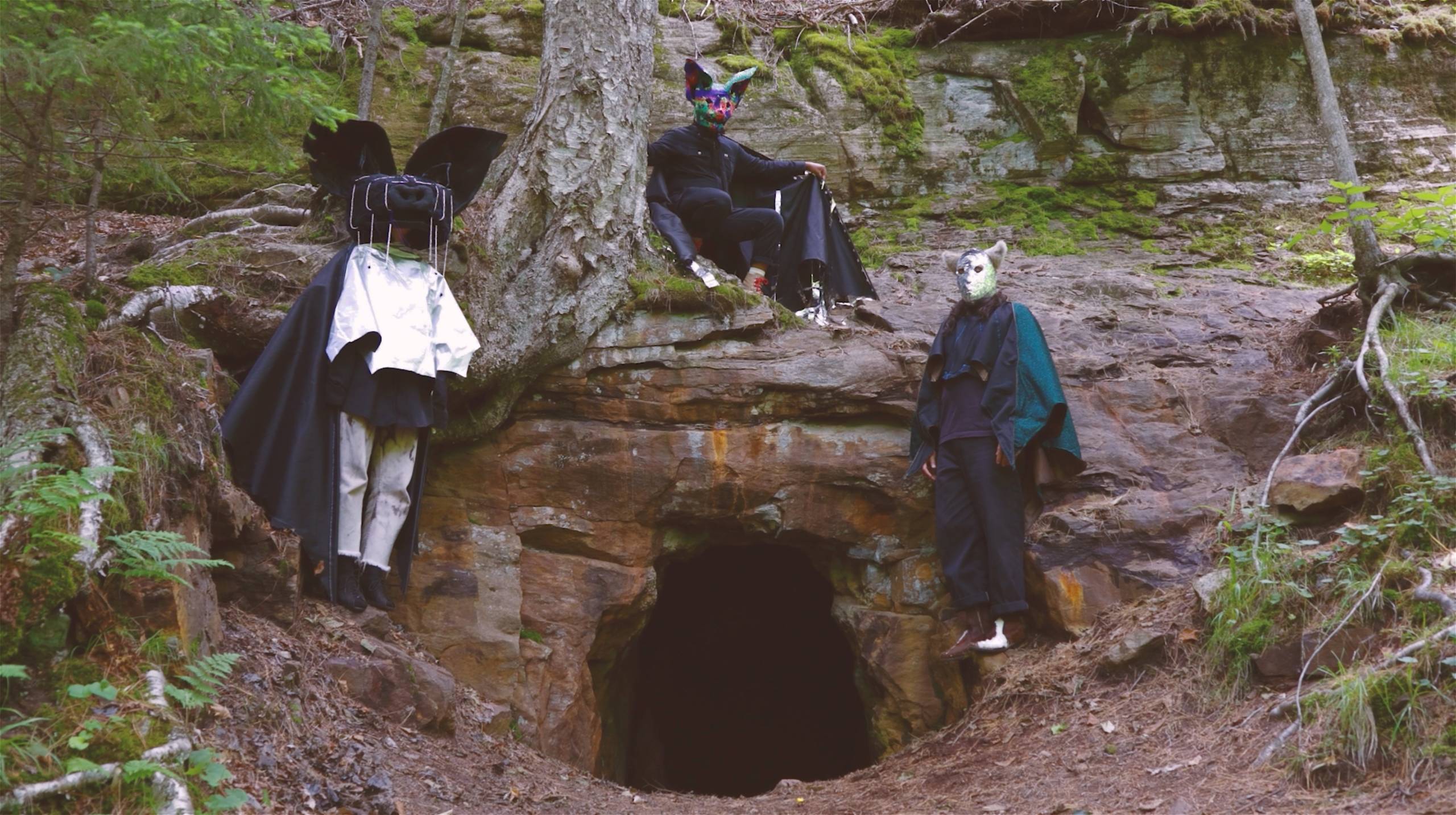 Three people wearing animal masks and robes near a cave entrance on a rocky hillside.