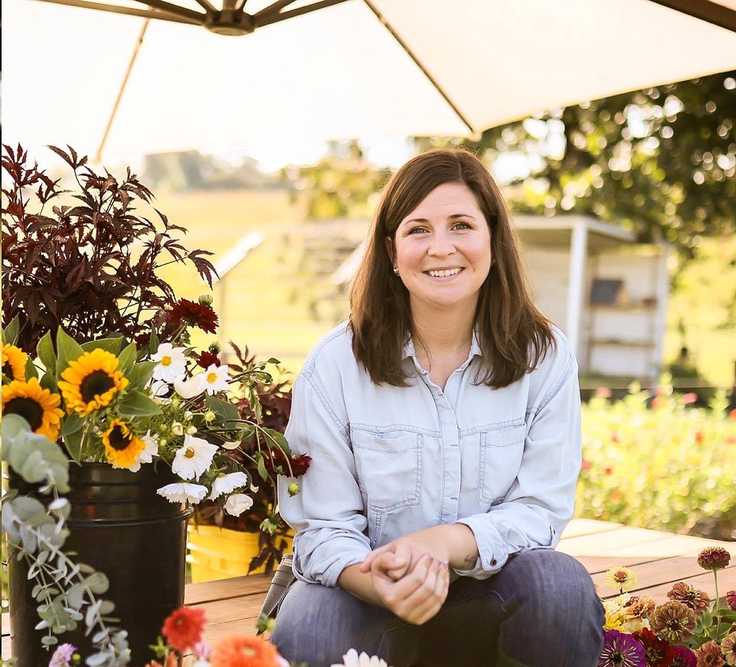 Woman in denim shirt and green boots sitting on bench surrounded by colorful flowers and umbrella outdoors.