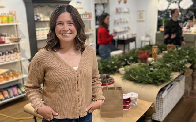 Woman in tan cardigan and jeans stands in a craft store with wreaths and supplies on tables.