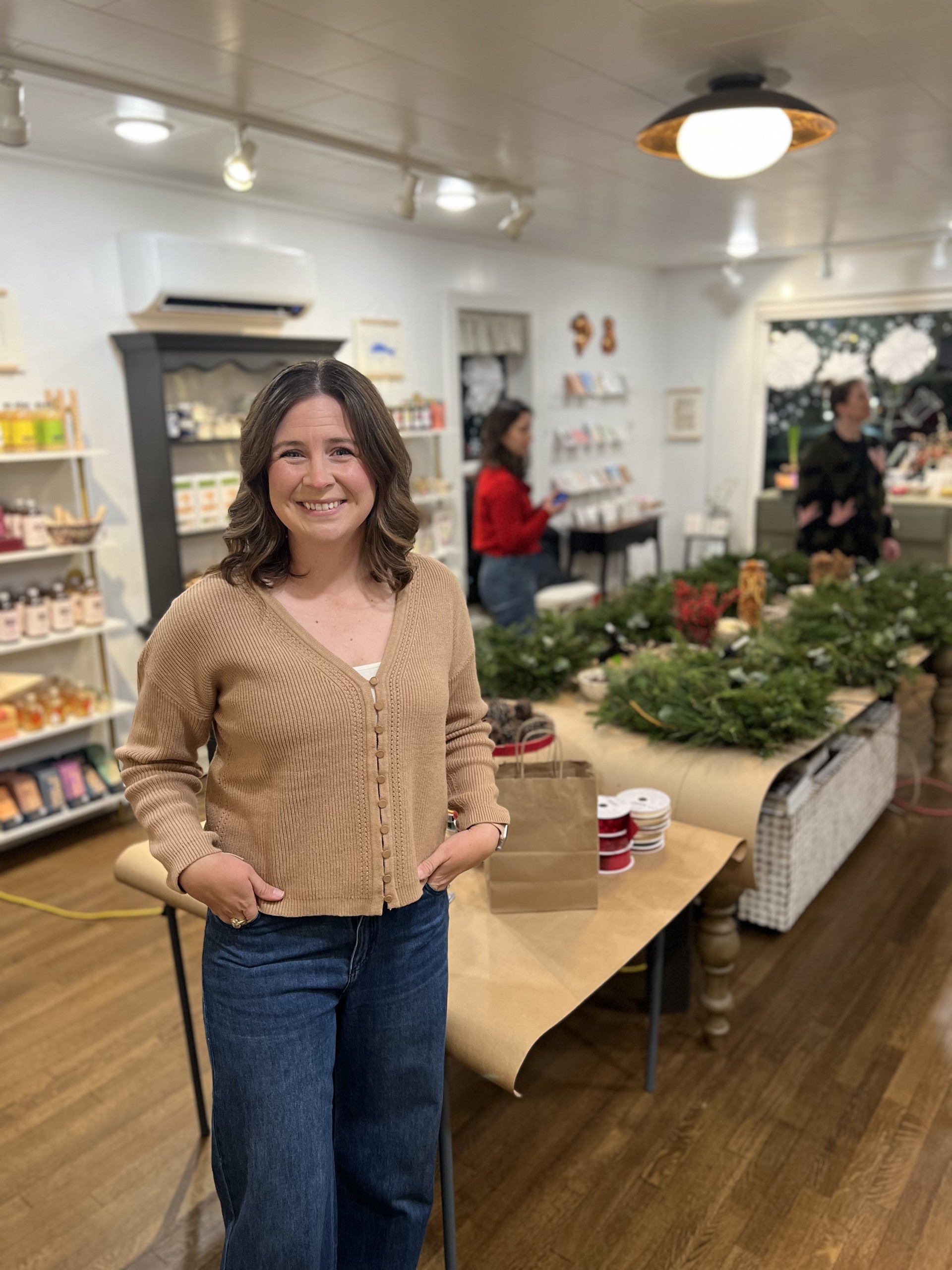 Woman in tan cardigan and jeans stands in a craft store with wreaths and supplies on tables.