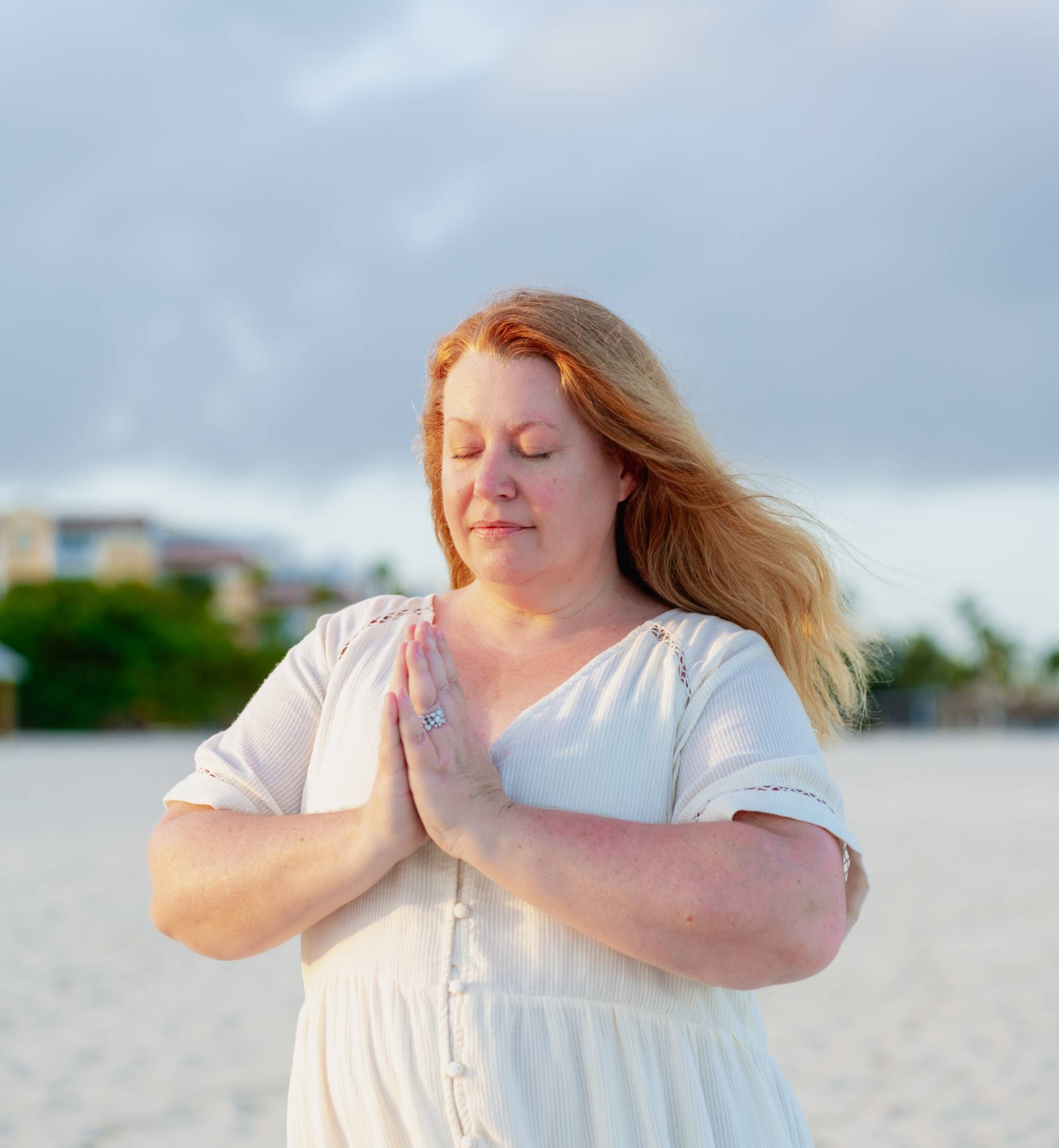 Woman in light dress meditating with hands pressed together on a sandy beach with buildings in background
