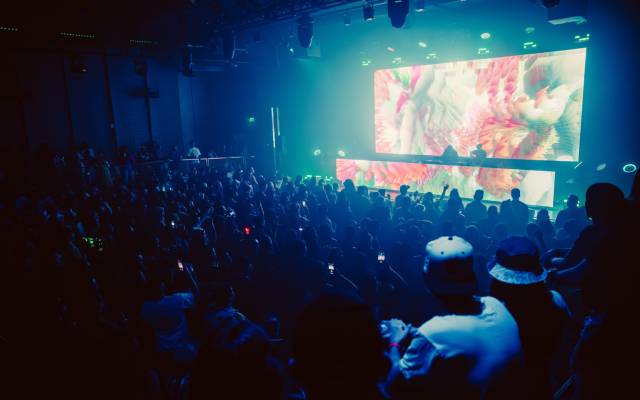 Indoor concert with a large crowd watching two DJs and colorful visuals on stage.