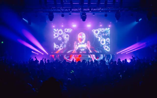 Crowded concert with two DJs on stage and vibrant digital visuals of a humanoid figure.