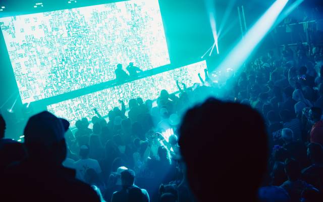 Large crowd at nightclub with two DJs performing on stage and bright digital screens.