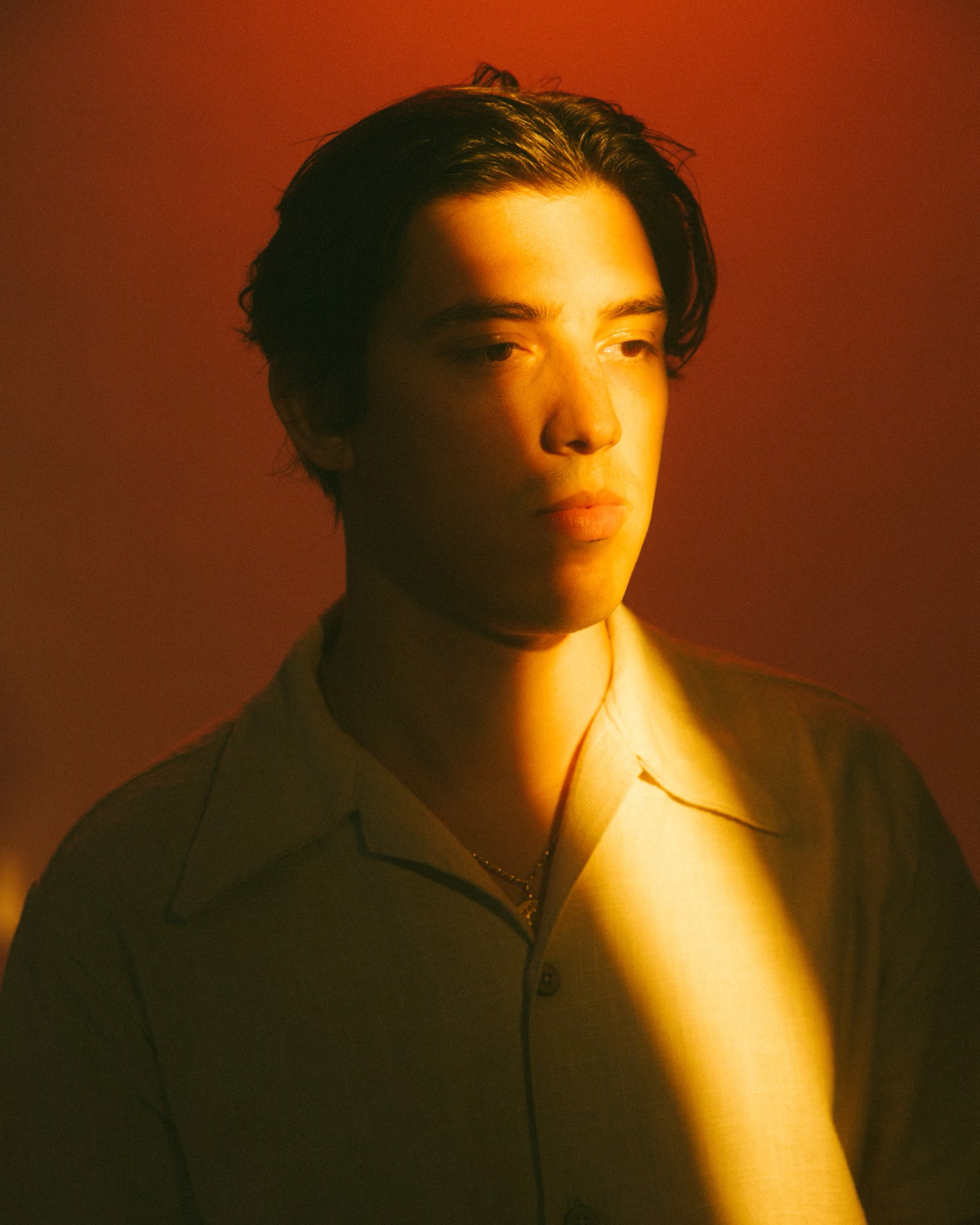 Young man in light shirt lit by a narrow warm beam against an orange background.