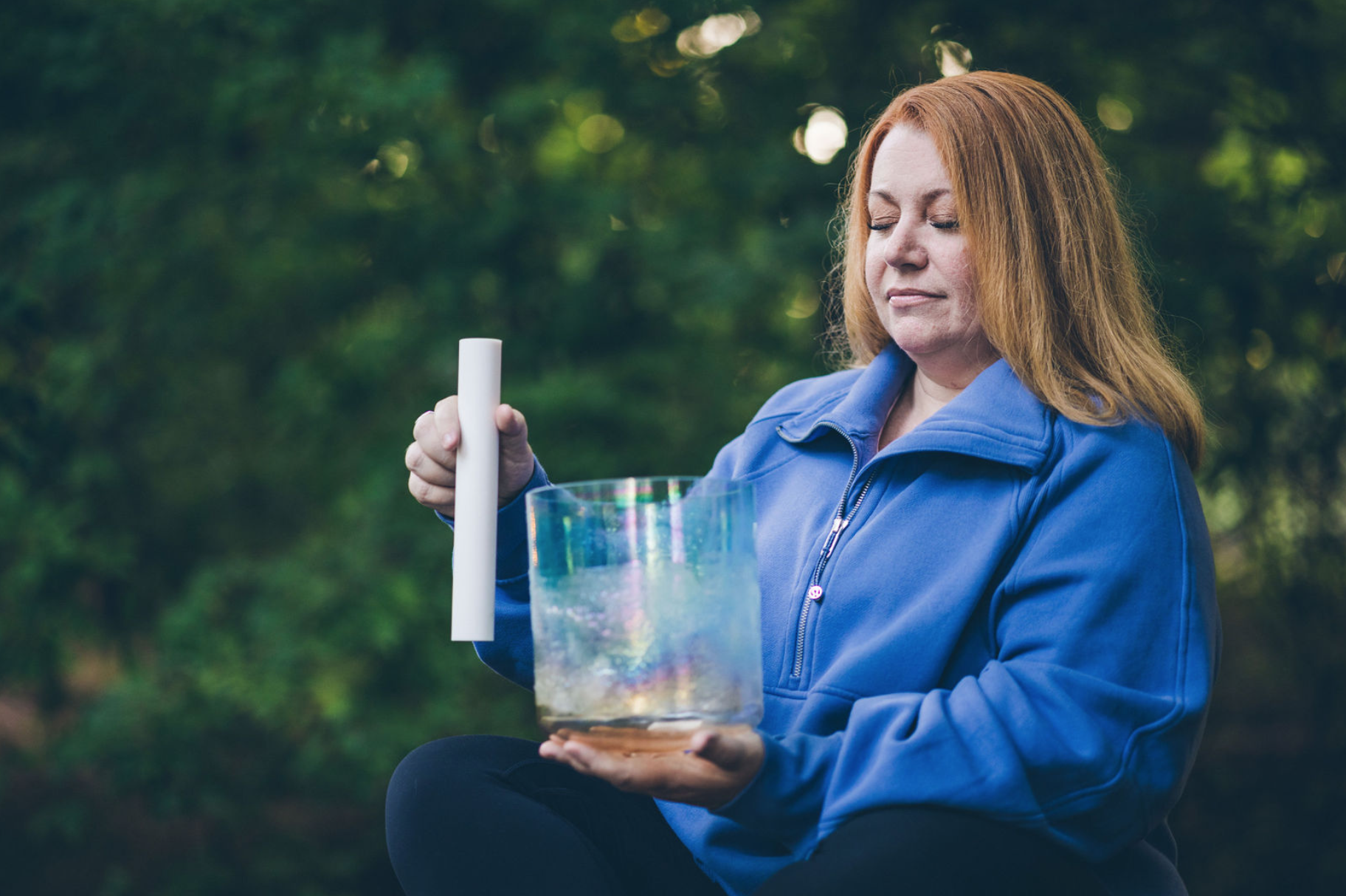 Woman in blue jacket holding a crystal singing bowl and mallet outdoors with eyes closed
