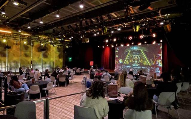 Audience seated at tables watching a live Oscars broadcast in a large dimly lit event space.