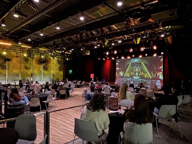 Audience seated at tables watching a live Oscars broadcast in a large dimly lit event space.