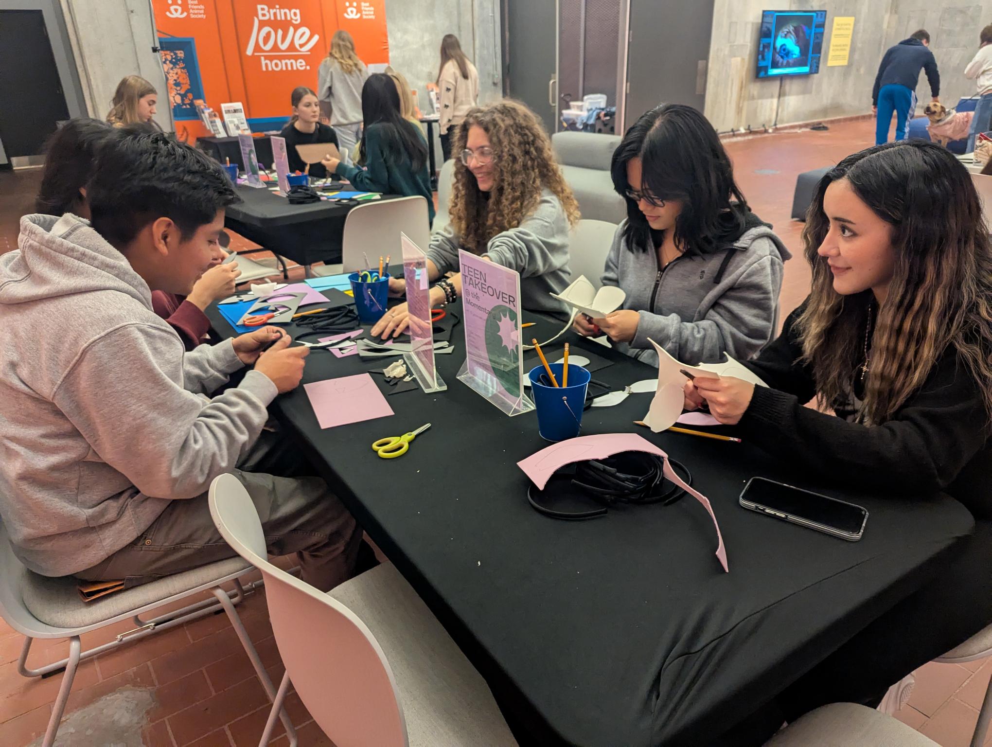 Teenagers seated at a table cutting paper and crafting in a community event space.