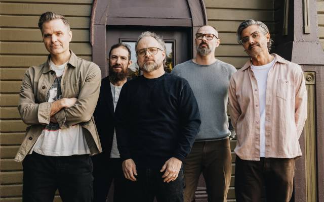 Five casually dressed men standing in front of a brown wooden building with a door.