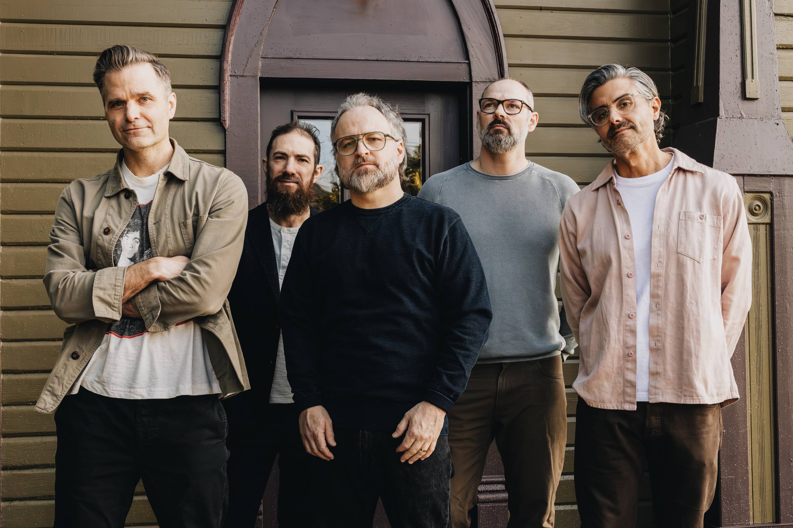 Five casually dressed men standing in front of a brown wooden building with a door.