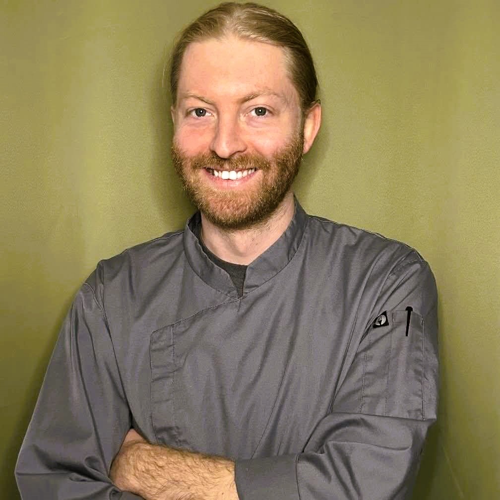 Light-skinned man with red hair and beard wearing a gray chef jacket against olive background.