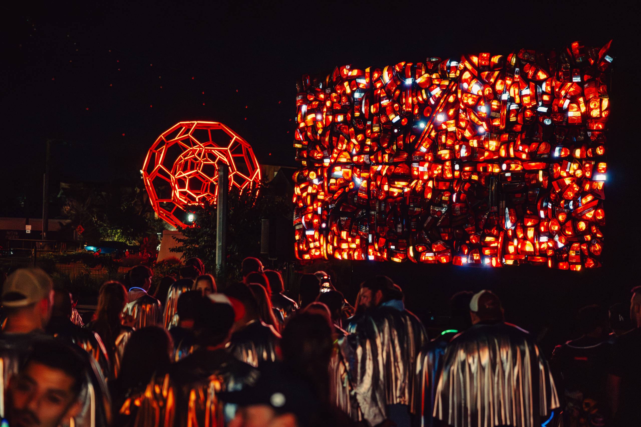 Crowd wearing silver capes viewing illuminated red geometric and rectangular light art installations at night.