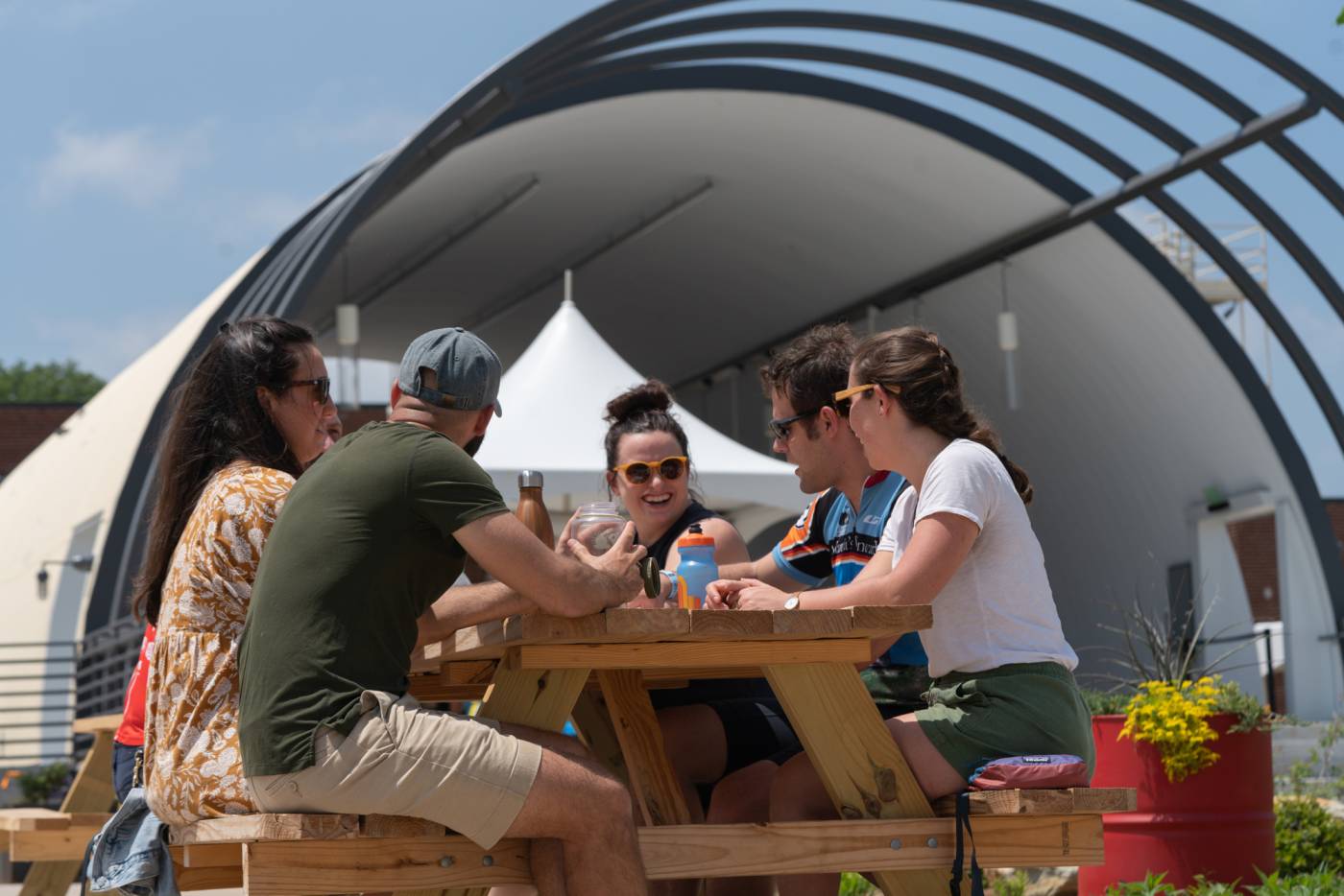 Five people sitting and talking at a wooden picnic table outdoors near a curved structure.