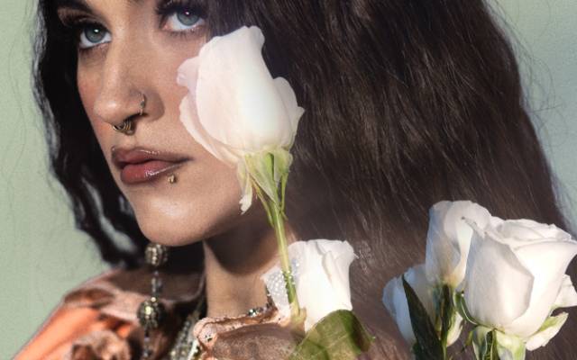 Woman with long dark hair and facial piercings holding white roses wearing orange clothing and jewelry