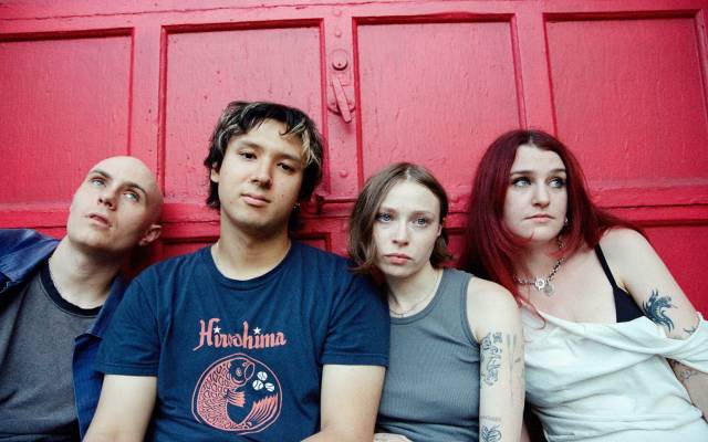 Four young people with tattoos sitting closely against a red wooden door with neutral expressions