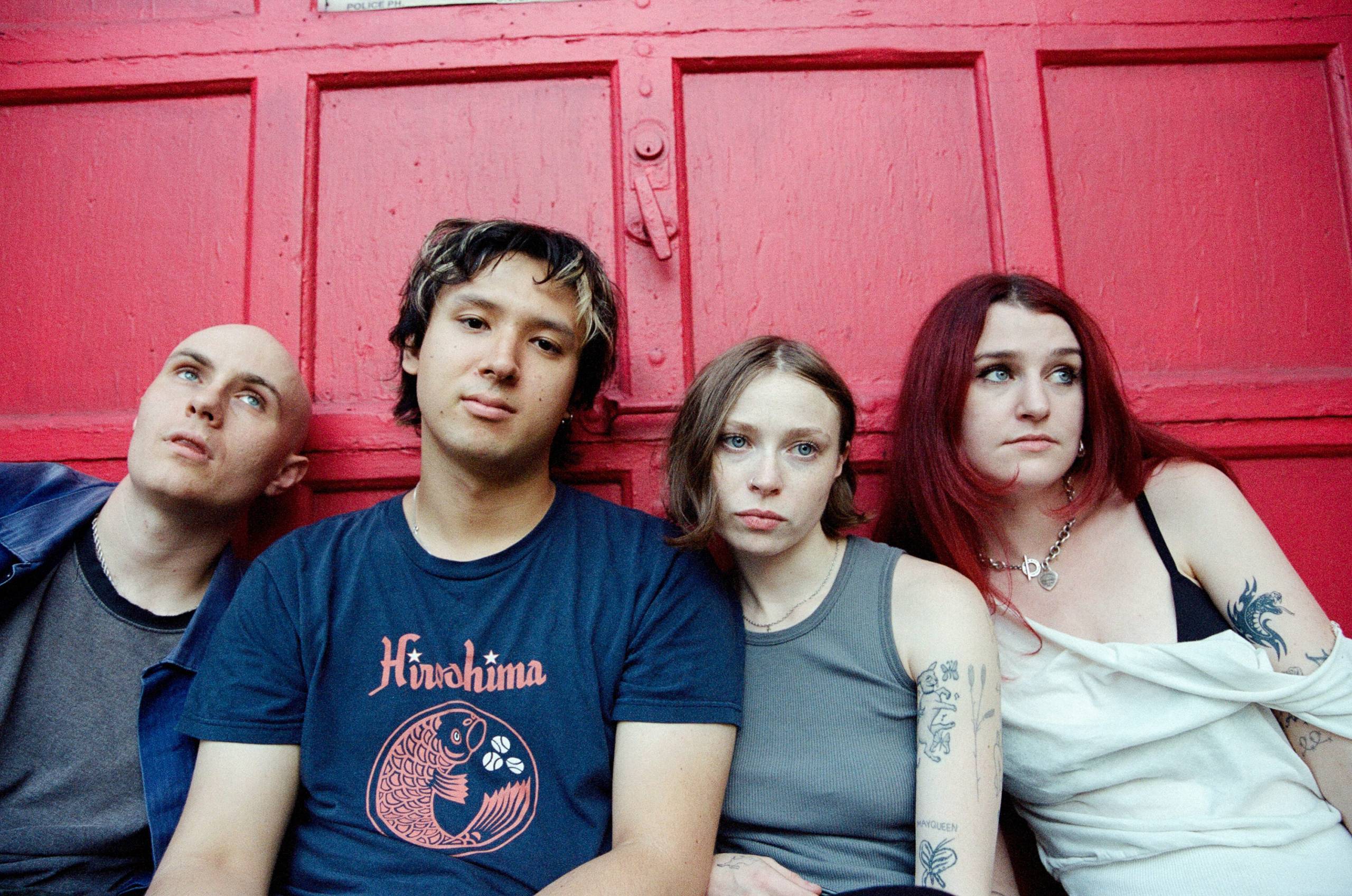 Four young people with tattoos sitting closely against a red wooden door with neutral expressions