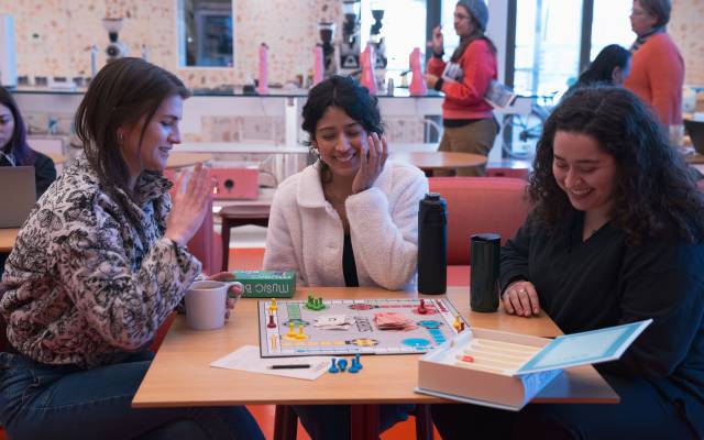 Three women sitting at a table in a casual setting playing a board game with game pieces and cards.