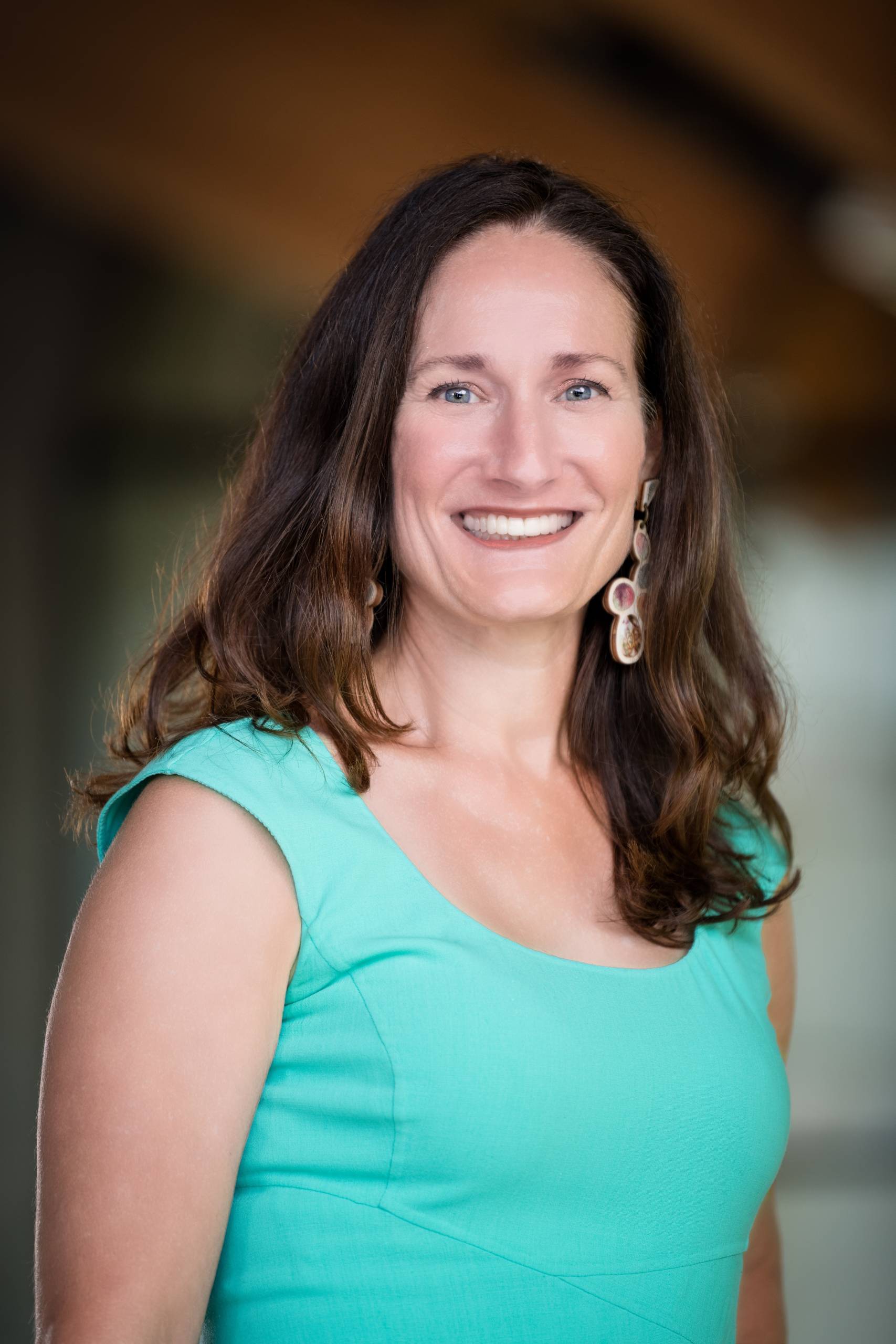 Woman with long brown hair wearing a sleeveless turquoise top and large earrings smiling at camera.