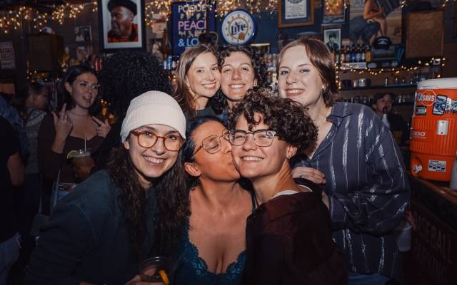 Six people smiling and posing together inside a dimly lit bar with string lights and decorations.