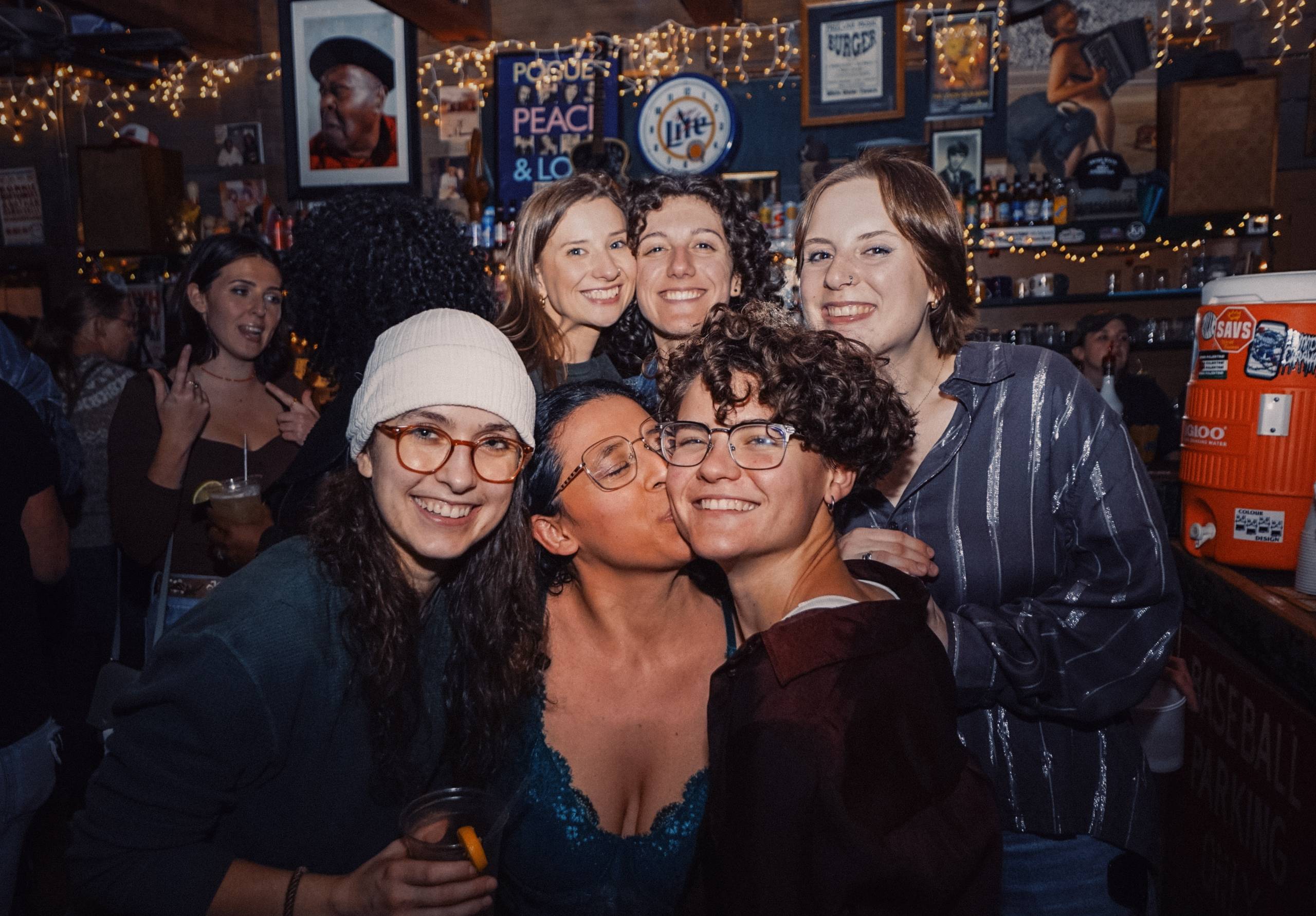 Six people smiling and posing together inside a dimly lit bar with string lights and decorations.