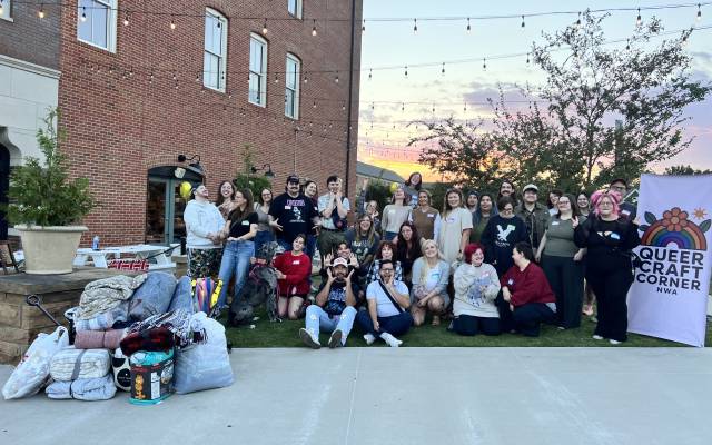 Group of people posing outdoors at sunset with blankets, a dog, and a Queer Craft Corner NWA banner.