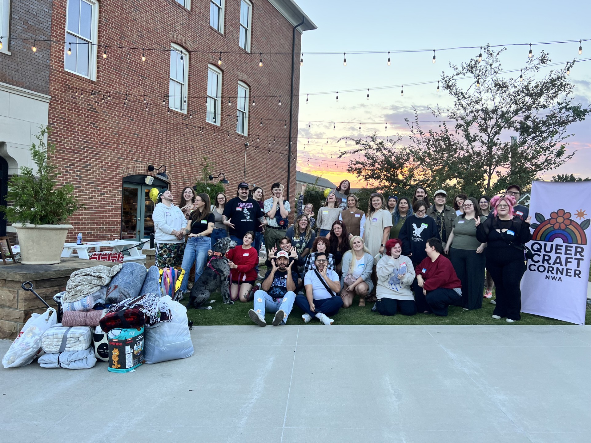 Group of people posing outdoors at sunset with blankets, a dog, and a Queer Craft Corner NWA banner.