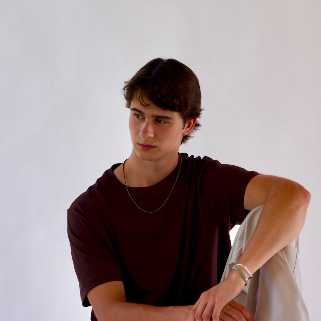 Young man sitting with one knee raised wearing a dark brown t-shirt, light pants, and jewelry.