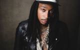 Man with dreadlocks wearing white and black hat, leather jacket, floral shirt, and chain necklace.