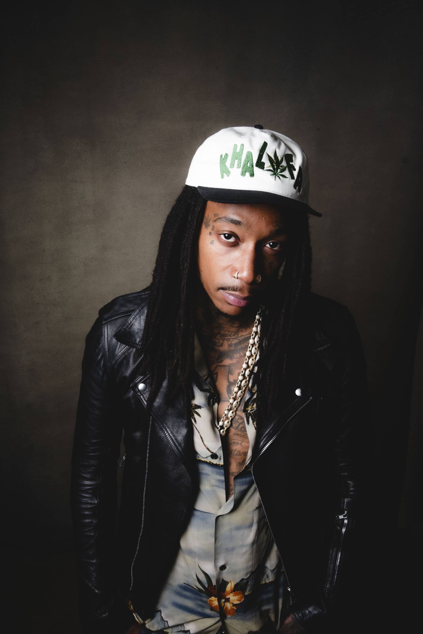 Man with dreadlocks wearing white and black hat, leather jacket, floral shirt, and chain necklace.