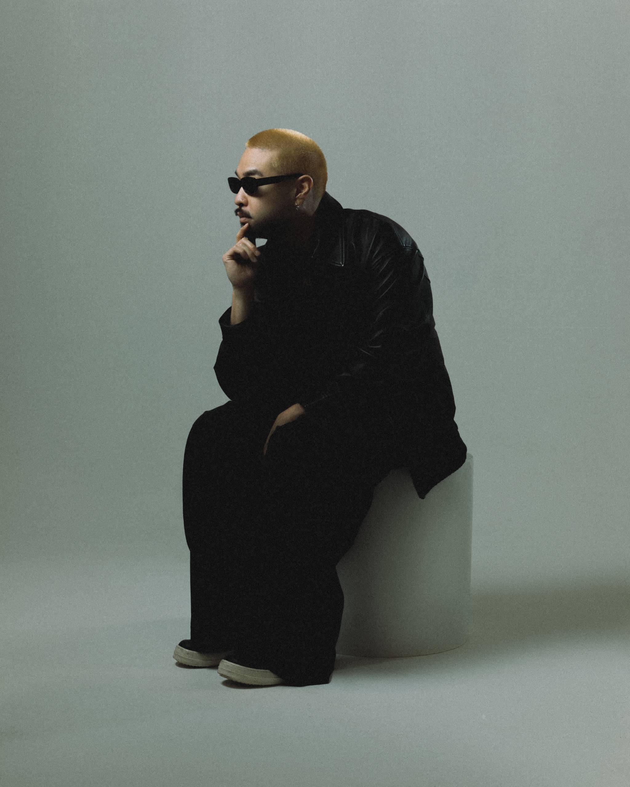 Person with short blonde hair wearing black jacket and sunglasses, seated on a white block against a gray backdrop.