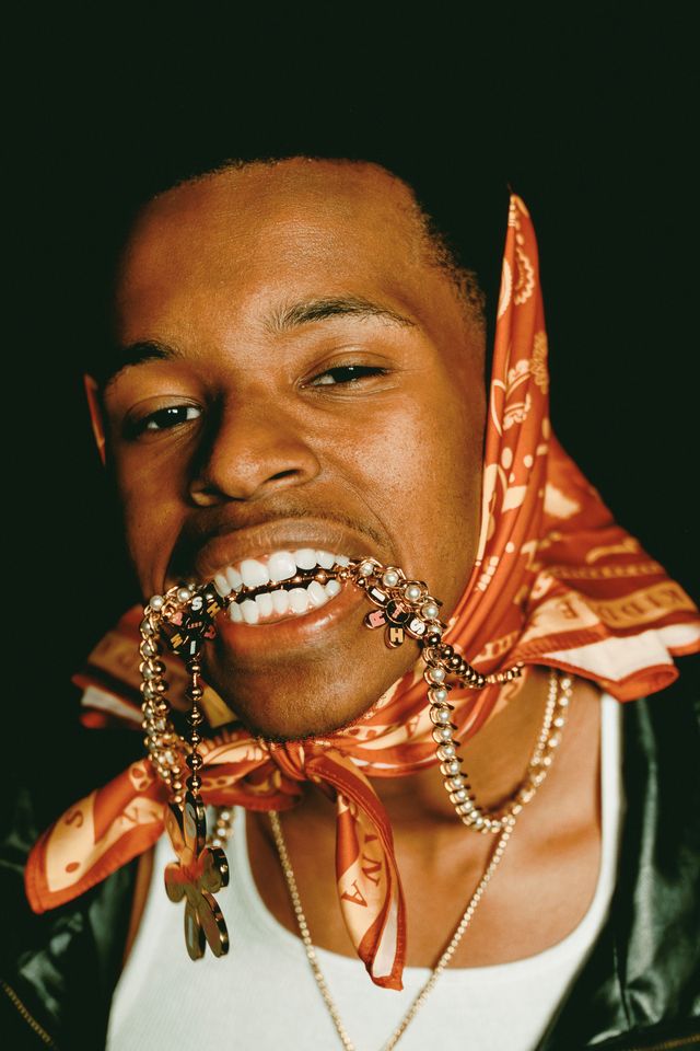 Young man wears red bandana, biting beaded bracelets, with a bright smile against a dark background.