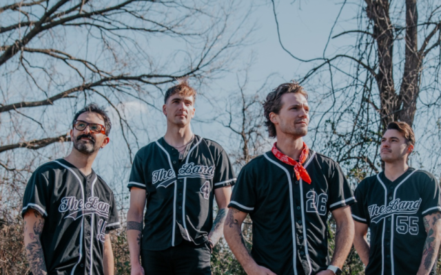 Four men in black "The Band" jerseys stand outdoors with leafless trees behind.