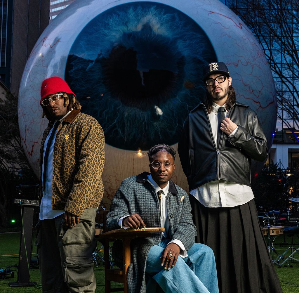 Three men posing in front of a giant illuminated eyeball sculpture outdoors.