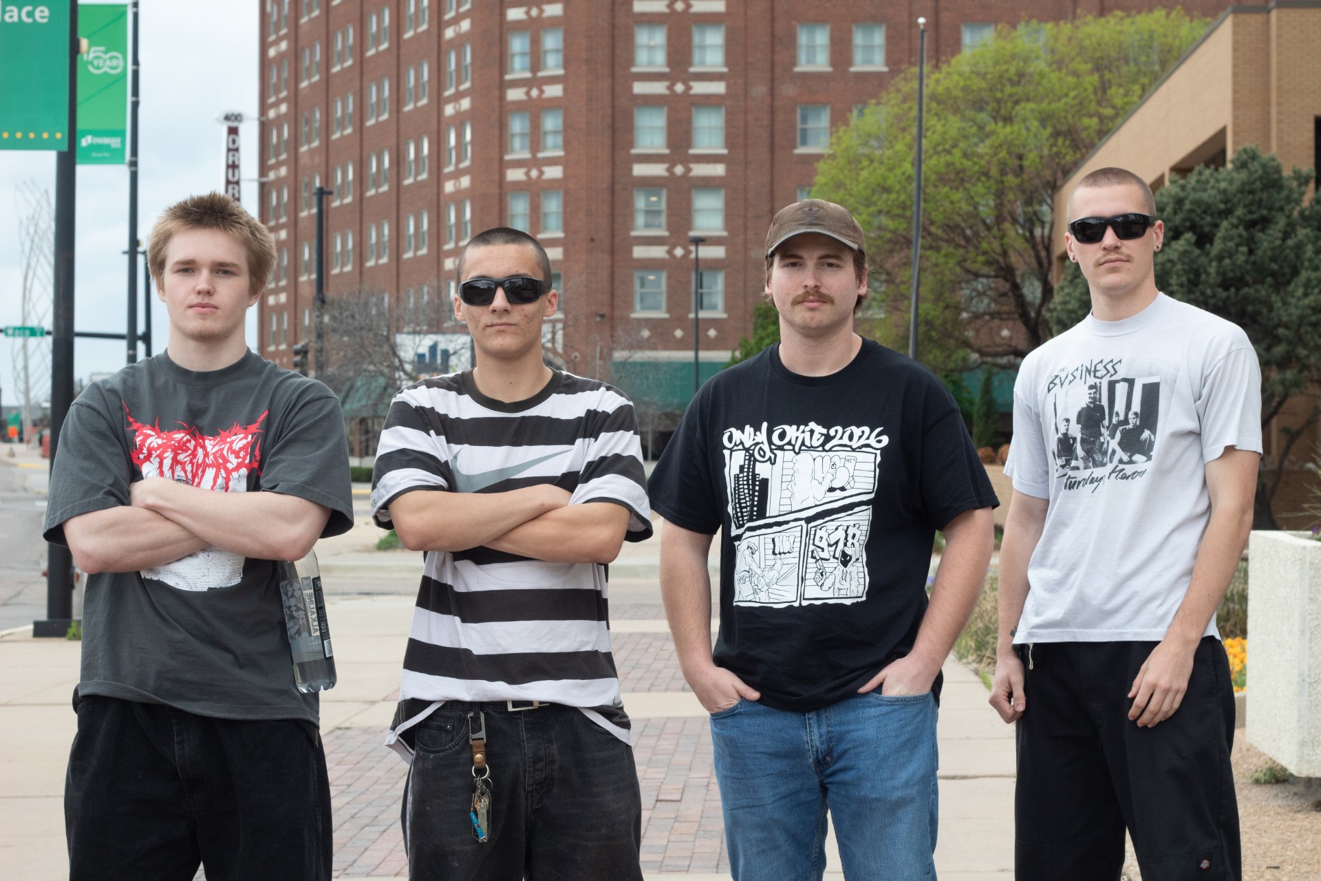 Four young men standing with arms crossed on city sidewalk, buildings in background.