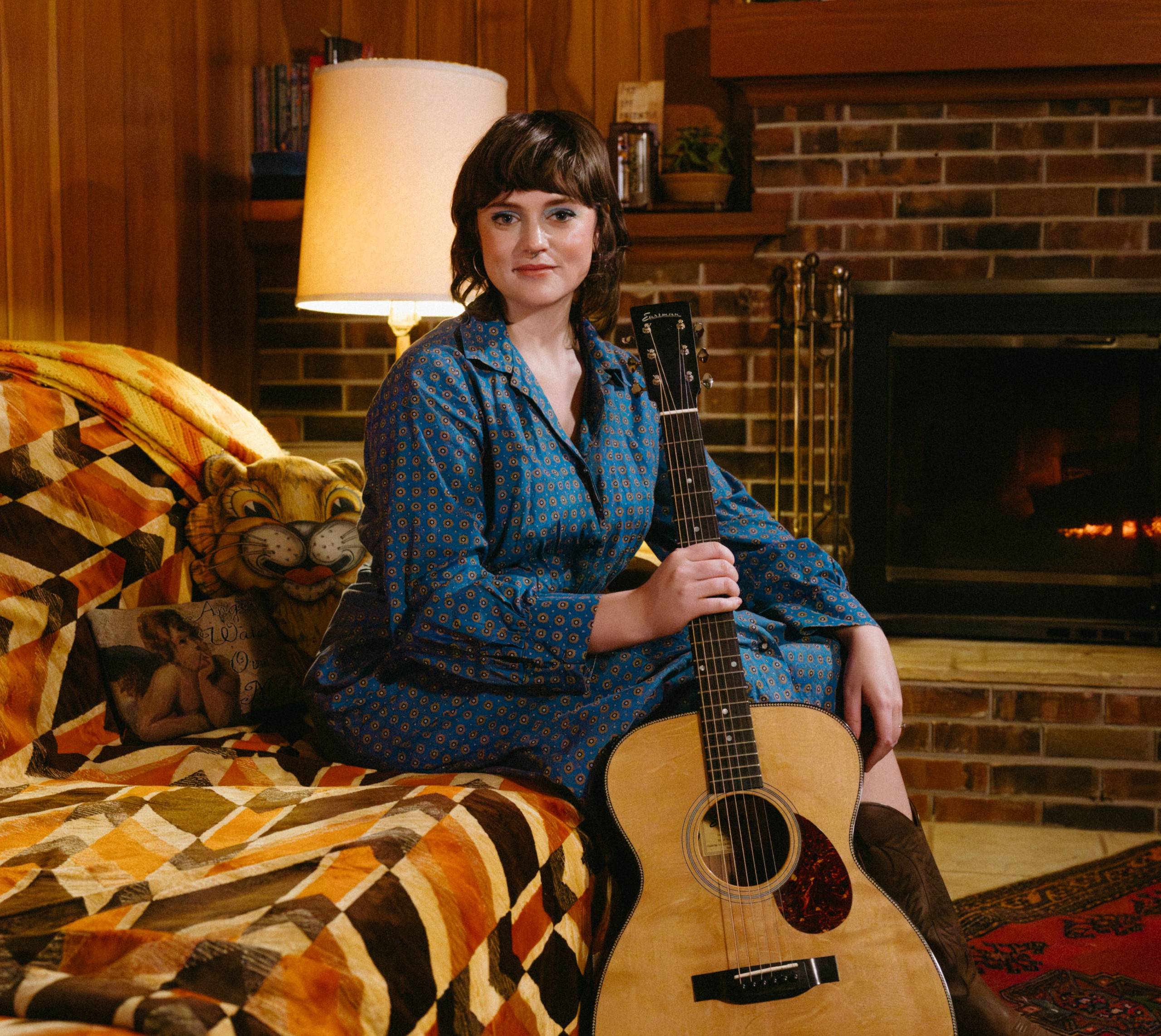 Woman in blue patterned dress sits with an acoustic guitar in a cozy room by a brick fireplace.