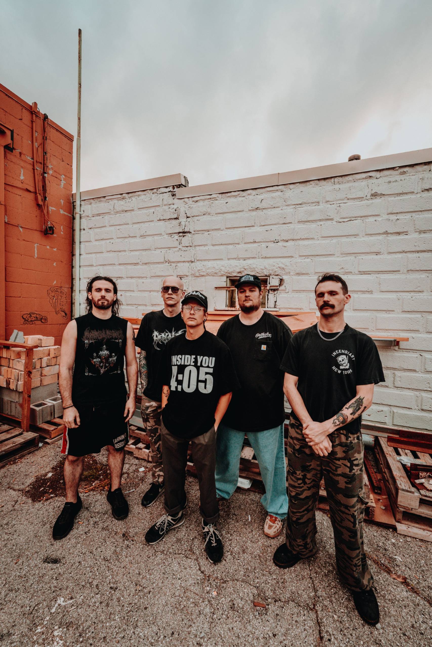 Five men pose in a concrete yard against white and orange walls with pallets nearby.