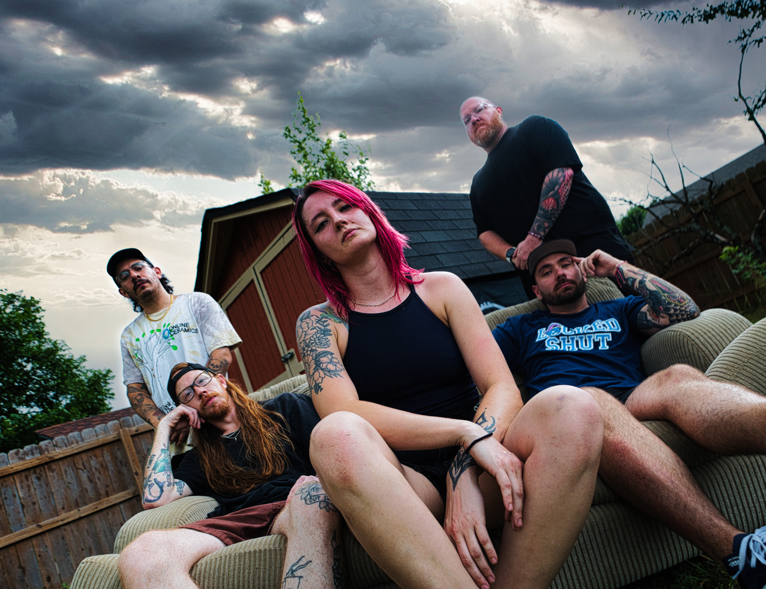 Group of five people with tattoos lounging on outdoor sofa in backyard under cloudy sky.