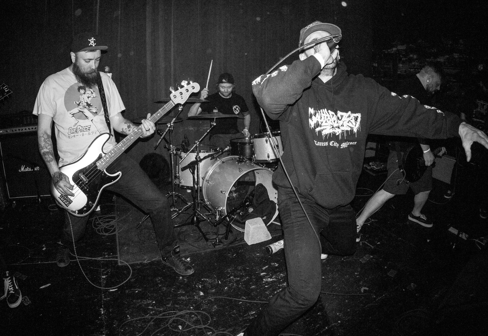 Black-and-white band performance with bass guitarist, drummer, and vocalist on a cluttered stage.