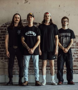 Four men stand side by side indoors against a brick wall, casual clothing and neutral expressions.
