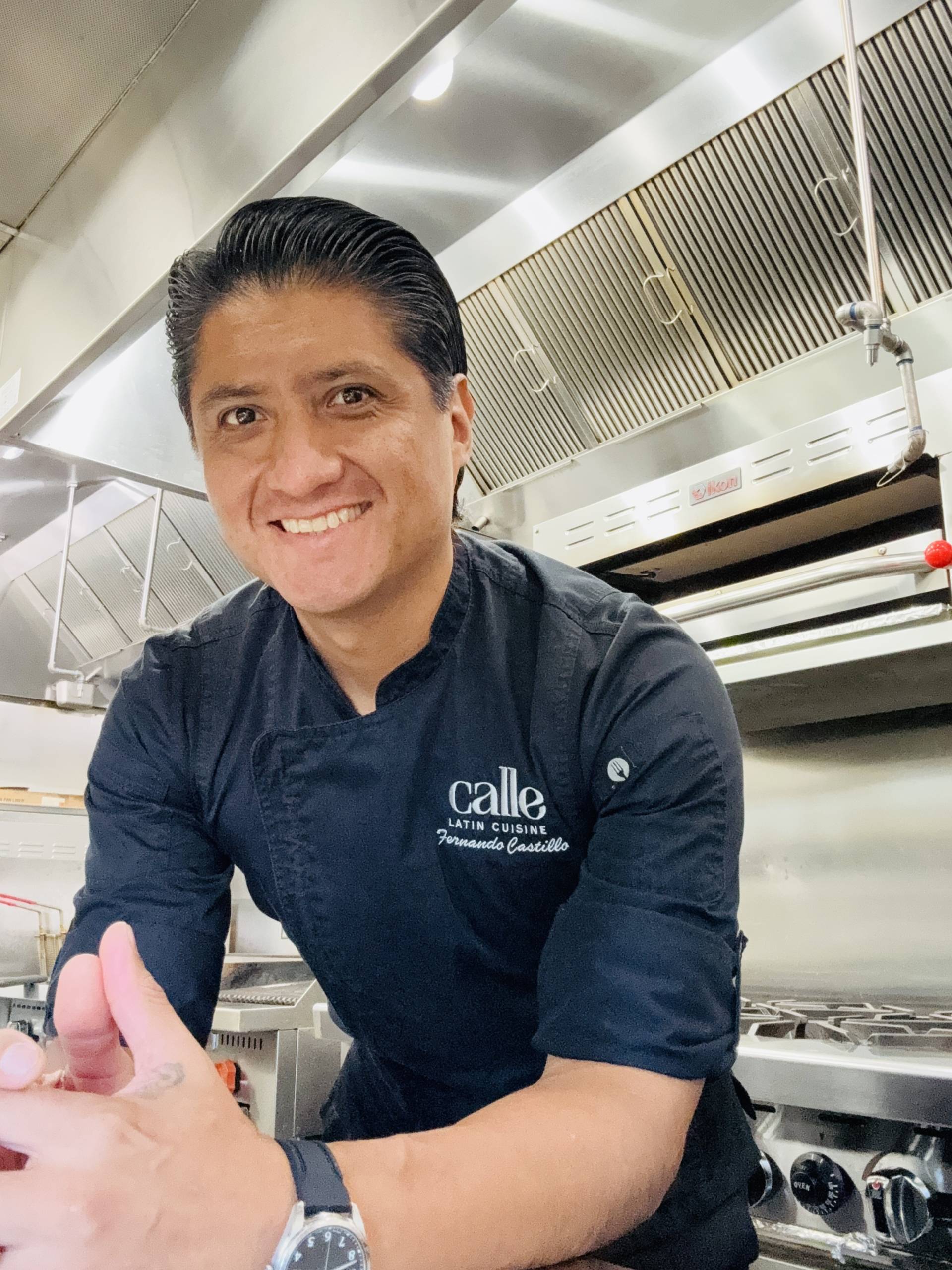 Smiling man in dark chef coat in a professional kitchen with stainless steel equipment behind him.
