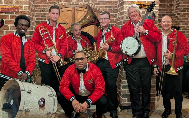 Group of six musicians in red jackets with brass instruments in a brick-walled setting.