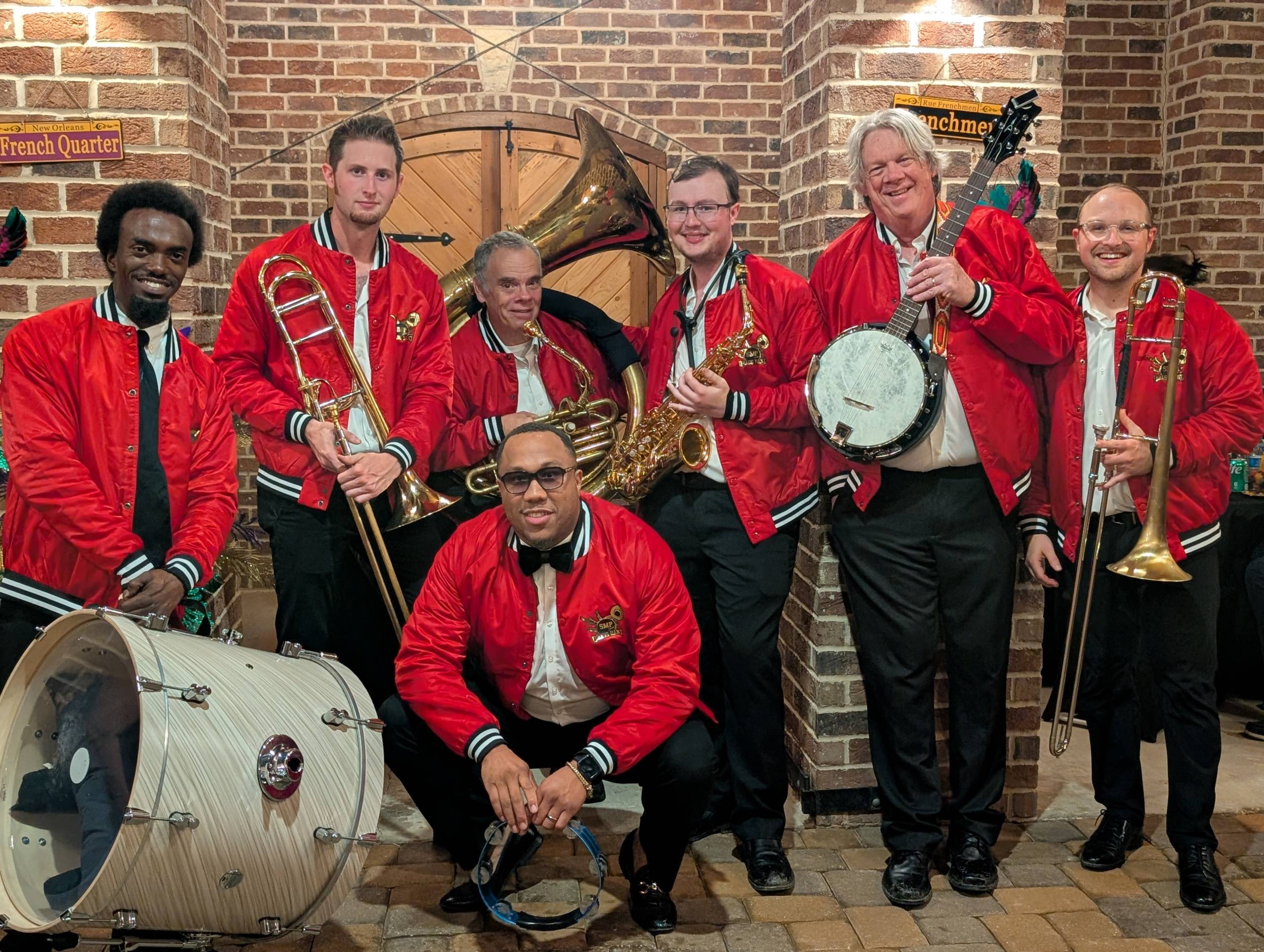 Group of six musicians in red jackets with brass instruments in a brick-walled setting.