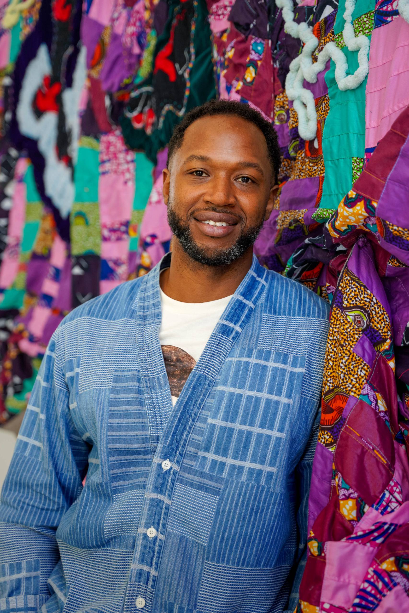 A smiling man with a beard wearing a blue plaid shirt stands in front of a colorful quilt backdrop.